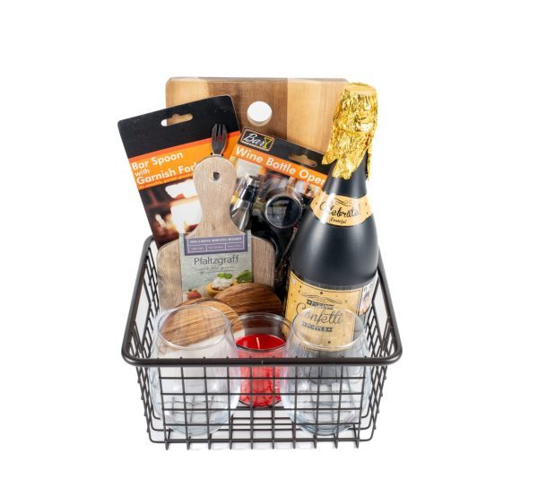 The Personalized Celebration Basket Medium with 2 Custom Coasters includes a metal wire basket that holds a bottle of sparkling wine, a wooden cutting board with a visible handle, a set of bar spoon and garnish fork, a wine bottle opener, a small wooden jar, two glass candles, and custom coasters. Stylish packaging labels are also part of the presentation.