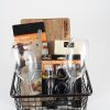 A black wire basket featuring the Personalized Celebration set, which includes a wine bottle opener set, two empty Riedel wine glasses, a bar spoon, a custom cutting board, and a box of assorted corks. The items are neatly arranged within the basket against a plain white background, ready for any personalized celebration.