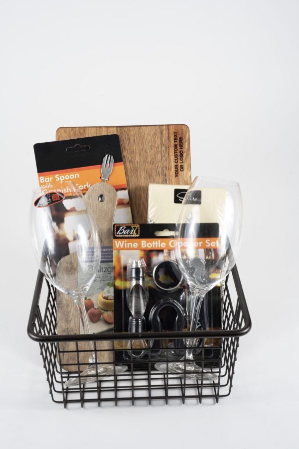 A black wire basket featuring the Personalized Celebration set, which includes a wine bottle opener set, two empty Riedel wine glasses, a bar spoon, a custom cutting board, and a box of assorted corks. The items are neatly arranged within the basket against a plain white background, ready for any personalized celebration.