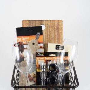 The Celebration Basket with 2 Stemmed Riedel Glasses - JUST ADD WINE includes various bar accessories such as two stemmed Riedel glasses, a wooden stirring spoon, a wooden cutting board, a bar spoon with a bottle opener, and a wine bottle opener set, all in their original packaging.