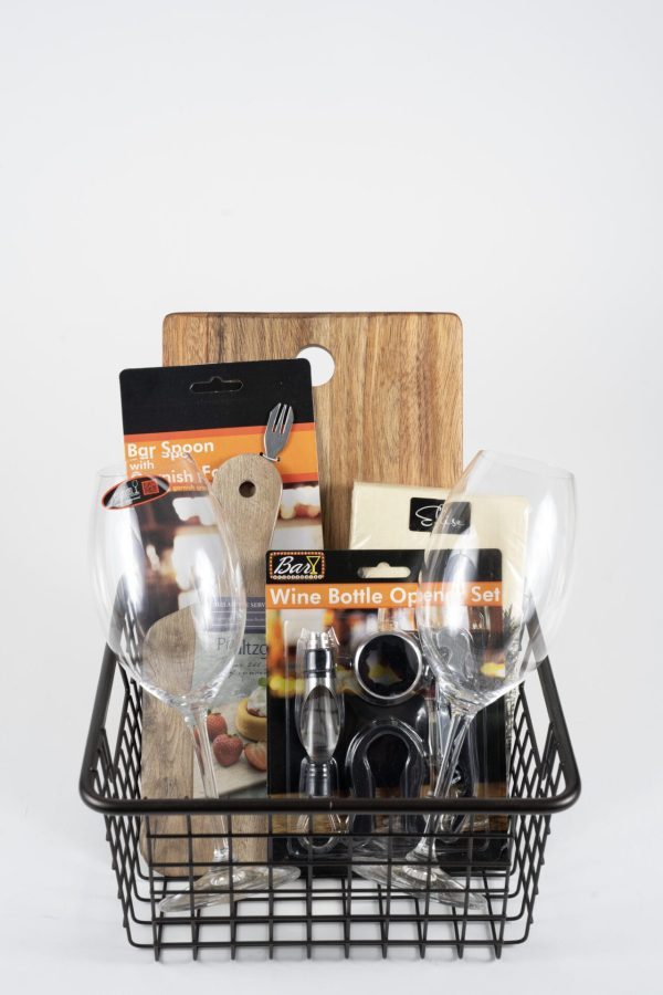 The Celebration Basket with 2 Stemmed Riedel Glasses - JUST ADD WINE includes various bar accessories such as two stemmed Riedel glasses, a wooden stirring spoon, a wooden cutting board, a bar spoon with a bottle opener, and a wine bottle opener set, all in their original packaging.