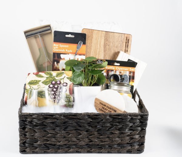 The Personalized Kitchen & Home Gift Basket features a variety of kitchen and bar items, including a bar spoon with a garnish fork, a cutting board set, two custom coasters, a plant, a grape-design kitchen towel, two custom glasses, and a jar.