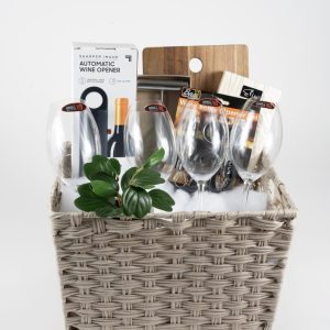 The Celebration Basket with 4 Riedel Glasses - JUST ADD WINE includes four empty Riedel glasses, a plant, an automatic wine opener, a wooden cutting board, a cheese slicer, and two packaged items with partially visible labels. The background is plain white.