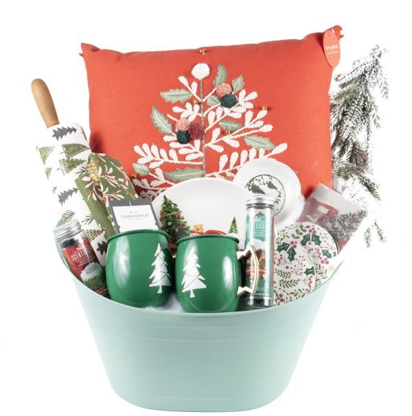 The "Christmas Home & Kitchen Gift Basket - Premium" includes a red pillow adorned with an embroidered Christmas tree, a rolling pin, two green mugs featuring white tree designs, pine-scented candles, a festive plate, and various other holiday-themed items, all beautifully arranged in a green bucket.