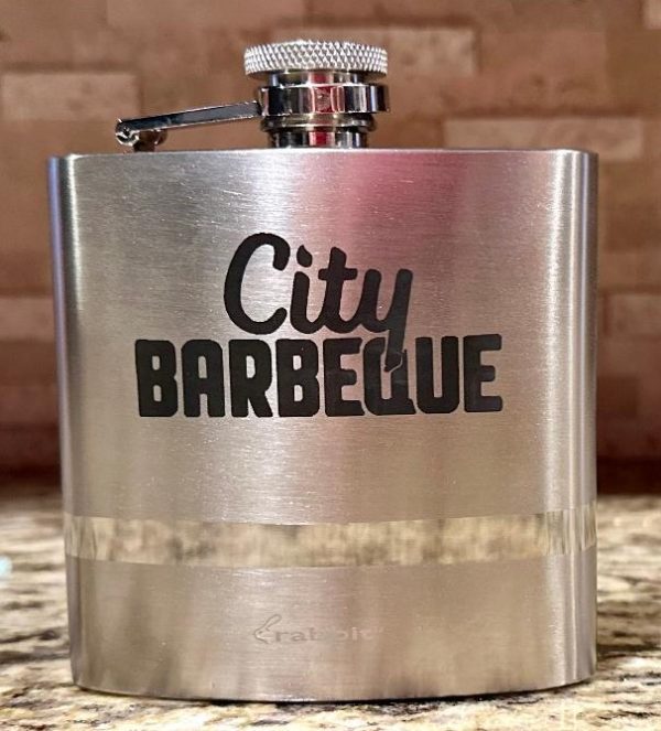 A sleek, silver flask with a screw-on cap rests on a speckled countertop. Adorned with the "City Barbeque" logo in bold, black lettering, this Silver Flask - Your Custom Text/Logo Here stands out. The background features a tiled wall.