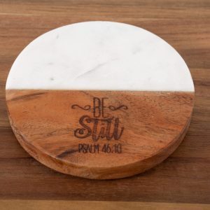 The "Be Still - Round" coaster, made of two materials—half white marble and half wood—sits gracefully on a wooden surface. The wooden portion is intricately engraved with the phrases "Be Still" and "Psalm 46:10" in decorative fonts, adding a touch of elegance to any setting.