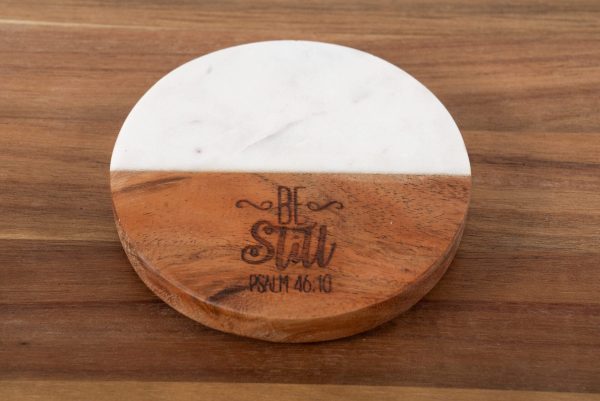 The "Be Still - Round" coaster, made of two materials—half white marble and half wood—sits gracefully on a wooden surface. The wooden portion is intricately engraved with the phrases "Be Still" and "Psalm 46:10" in decorative fonts, adding a touch of elegance to any setting.