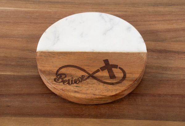 A round coaster named "Believe - Round" sitting on a wooden surface. The coaster features a design that is half white marble and half brown wood, with an infinity symbol and cross engraved on the wooden part. The word "Believe" is stylishly integrated into the infinity symbol.