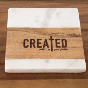 The "Created with a Purpose - Square" coaster is displayed, showcasing a striking combination of white marble and brown wood. An earthy wooden stripe runs across the center, carrying the inscription "CREATED with a Purpose," with the letter "T" artistically fashioned as a cross. The coaster rests elegantly on a wooden surface.