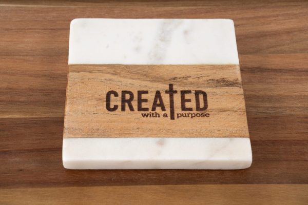 The "Created with a Purpose - Square" coaster is displayed, showcasing a striking combination of white marble and brown wood. An earthy wooden stripe runs across the center, carrying the inscription "CREATED with a Purpose," with the letter "T" artistically fashioned as a cross. The coaster rests elegantly on a wooden surface.