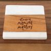 The "Every Moment Matters - Square" coaster boasts a white marble top and bottom with a wooden center, elegantly inscribed with the phrase "Every Moment Matters" in artistic black lettering. It is displayed on a wooden surface.