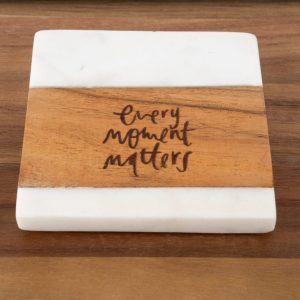 The "Every Moment Matters - Square" coaster boasts a white marble top and bottom with a wooden center, elegantly inscribed with the phrase "Every Moment Matters" in artistic black lettering. It is displayed on a wooden surface.