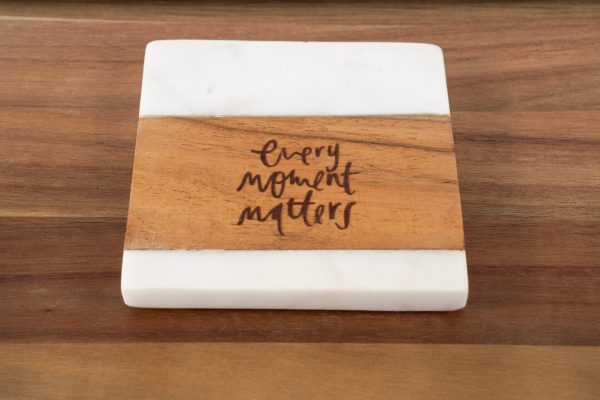 The "Every Moment Matters - Square" coaster boasts a white marble top and bottom with a wooden center, elegantly inscribed with the phrase "Every Moment Matters" in artistic black lettering. It is displayed on a wooden surface.