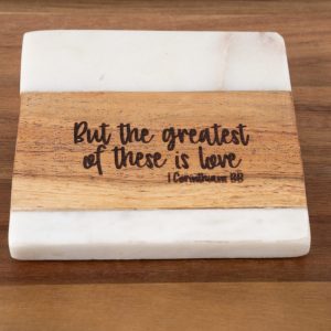 The "But the Greatest of these is Love - Square" plaque features a white top and bottom, with a wooden middle section inscribed with the text: "But the greatest of these is love 1 Corinthians 13:13." The plaque is elegantly displayed on a wooden surface.