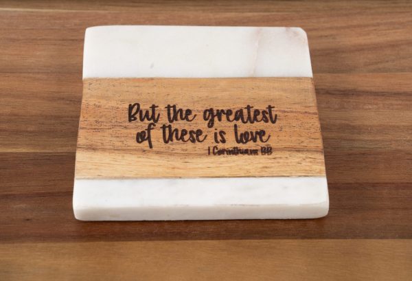 The "But the Greatest of these is Love - Square" plaque features a white top and bottom, with a wooden middle section inscribed with the text: "But the greatest of these is love 1 Corinthians 13:13." The plaque is elegantly displayed on a wooden surface.