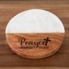 The "Prayer Changes Things - Round" coaster, featuring a two-tone design with a marble top half and a wooden bottom half, rests on a wooden surface. The wooden portion is engraved with the words "Prayer changes things," underscoring the transformative power of faith, and includes a small cross.