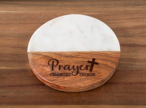 The "Prayer Changes Things - Round" coaster, featuring a two-tone design with a marble top half and a wooden bottom half, rests on a wooden surface. The wooden portion is engraved with the words "Prayer changes things," underscoring the transformative power of faith, and includes a small cross.