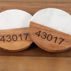 A pair of "Pair Round Coasters - Choose any Zip Code," featuring a blend of white marble and natural wood, sits side by side on a wooden surface. Each coaster is engraved with the zip code "43017" on the wooden part.