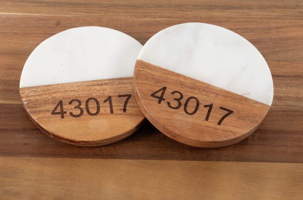 A pair of "Pair Round Coasters - Choose any Zip Code," featuring a blend of white marble and natural wood, sits side by side on a wooden surface. Each coaster is engraved with the zip code "43017" on the wooden part.