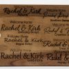 A wooden board with a hole on the left side showcases various text styles for "Welcome Home Rachel & Kirk." The displayed fonts include Allura, Dancing Script, Segoe Script, Gabriola, Southam Demo, Great Vibes, and Honey Script. It's an ideal addition to the Personalized Kitchen & Home Gift Basket with 2 Custom Glasses, 2 Custom Coasters & Cutting Board.