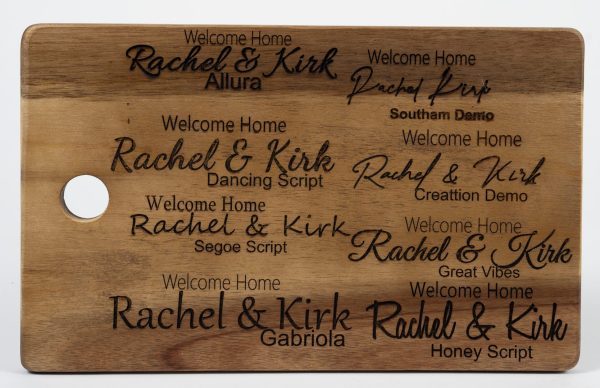 A wooden board with a hole on the left side showcases various text styles for "Welcome Home Rachel & Kirk." The displayed fonts include Allura, Dancing Script, Segoe Script, Gabriola, Southam Demo, Great Vibes, and Honey Script. It's an ideal addition to the Personalized Kitchen & Home Gift Basket with 2 Custom Glasses, 2 Custom Coasters & Cutting Board.