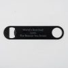 A flat black bottle opener, etched on both sides with the inscription, "World's Best Dad! Love, The Reason You Drink!" The opener features a circular hole on one end and a flat bottle opening mechanism on the other. The background is plain white.