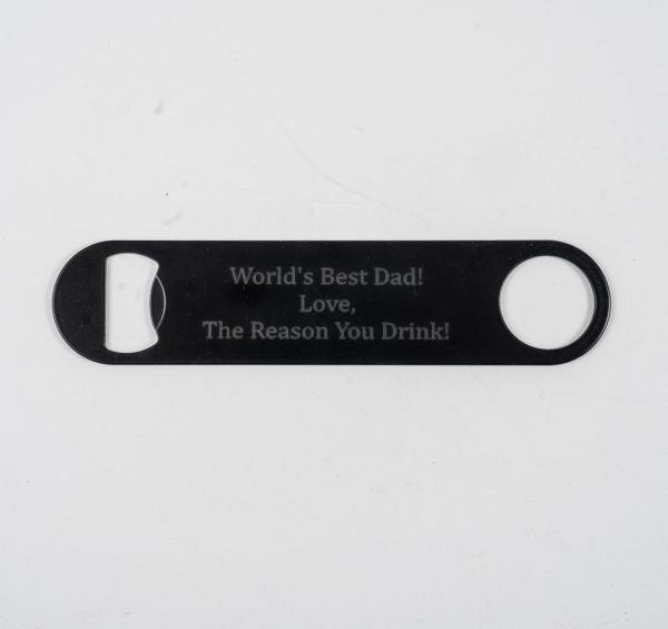 A flat black bottle opener, etched on both sides with the inscription, "World's Best Dad! Love, The Reason You Drink!" The opener features a circular hole on one end and a flat bottle opening mechanism on the other. The background is plain white.