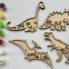 The Dinosaur Craft Kit is displayed on a white background. It includes wooden cutouts of a Brachiosaurus, Triceratops, T-Rex, and Pterodactyl adorned with palm tree designs. The kit also comes with five paintbrushes, four paint colors in pods, and a container of glue.