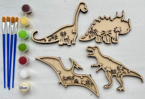 The Dinosaur Craft Kit is displayed on a white background. It includes wooden cutouts of a Brachiosaurus, Triceratops, T-Rex, and Pterodactyl adorned with palm tree designs. The kit also comes with five paintbrushes, four paint colors in pods, and a container of glue.