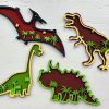 Four colorful, wooden dinosaurs against a white background. From the top left clockwise: a pterodactyl with red details and star cutouts, a T-rex, a triceratops adorned with green leaves, and a long-necked herbivore that could be a brachiosaurus. This Dinosaur Craft Kit is perfect for any craft project or dinosaur enthusiast!