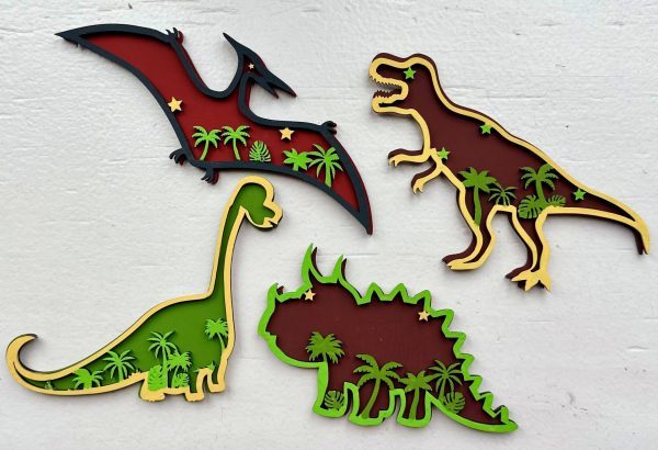 Four colorful, wooden dinosaurs against a white background. From the top left clockwise: a pterodactyl with red details and star cutouts, a T-rex, a triceratops adorned with green leaves, and a long-necked herbivore that could be a brachiosaurus. This Dinosaur Craft Kit is perfect for any craft project or dinosaur enthusiast!