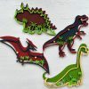 The Dinosaur Craft Kit includes wooden wall decorations depicting a Stegosaurus, Tyrannosaurus Rex, Pterodactyl, and Brachiosaurus in various colors. Each dinosaur is surrounded by stars and palm trees with contrasting color accents set against a white background.