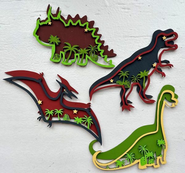 The Dinosaur Craft Kit includes wooden wall decorations depicting a Stegosaurus, Tyrannosaurus Rex, Pterodactyl, and Brachiosaurus in various colors. Each dinosaur is surrounded by stars and palm trees with contrasting color accents set against a white background.