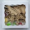 The Dinosaur Craft Kit includes wooden dinosaur cutouts and small plastic pots of paint in vibrant colors like red, green, yellow, and white. The packaging features a white frame with a transparent window that displays the dinosaur-themed contents inside.