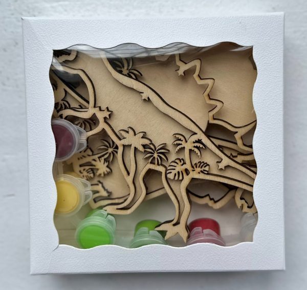 The Dinosaur Craft Kit includes wooden dinosaur cutouts and small plastic pots of paint in vibrant colors like red, green, yellow, and white. The packaging features a white frame with a transparent window that displays the dinosaur-themed contents inside.