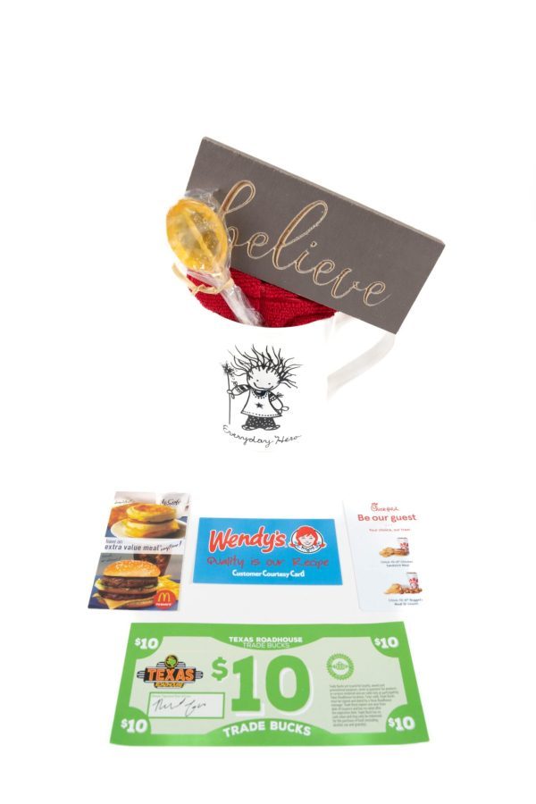 The Everyday Hero Mug Gift Basket with Free Food Certificate includes a white cup featuring a graphic of a person and the text "Everyday Hero." Inside the basket, you'll find a lollipop, a wooden sign that says "believe," and red fabric. Additionally, it comes with free food certificates and coupons from McDonald's, Wendy's, and Texas Roadhouse.