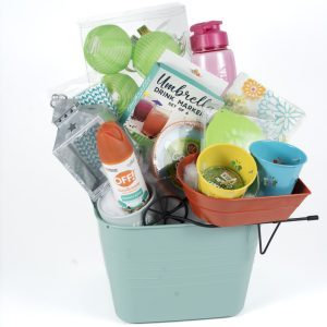 The Adult Gambling Night Basket is filled with assorted items, including a pink water bottle, green and blue paper lanterns, OFF! insect repellent, a lantern-shaped candle holder, gardening gloves, small flower pots, a yard game set, and a package labeled "Umbrella Drink Markers.