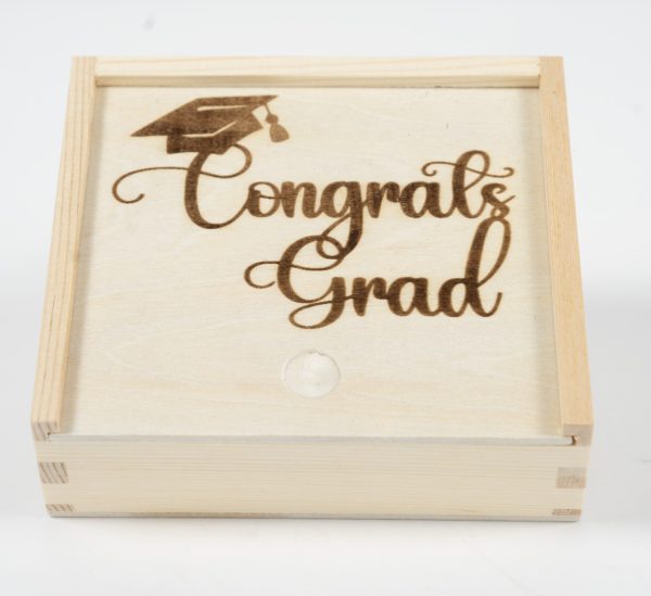 The "Gift Box - Congratulations Grad" is a wooden gift box featuring a sliding lid engraved with the words "Congrats Grad" beneath an intricately detailed graduation cap. This elegant box, ideal for celebrating such a significant achievement, is showcased against a pristine white background.