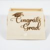 The "Gift Box - Congratulations Grad" is a small wooden box with an open lid that reveals a simple, empty interior. The lid features the words "Congratulations Grad" and a graduation cap graphic artistically burned into the wood.