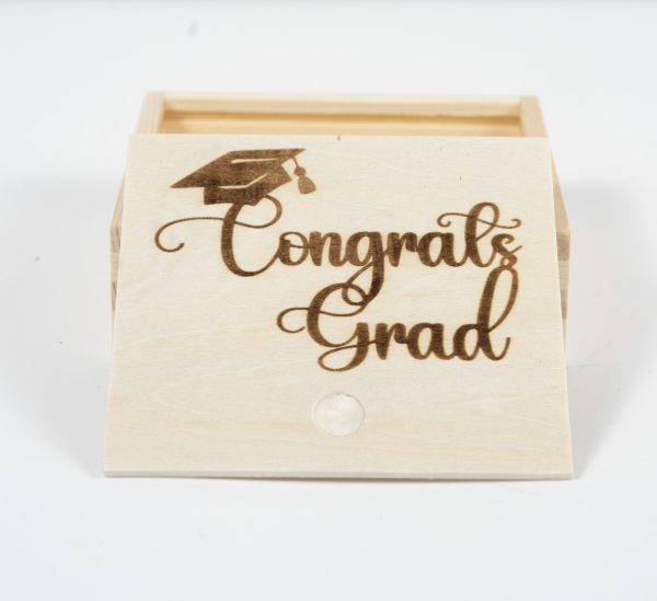 The "Gift Box - Congratulations Grad" is a small wooden box with an open lid that reveals a simple, empty interior. The lid features the words "Congratulations Grad" and a graduation cap graphic artistically burned into the wood.