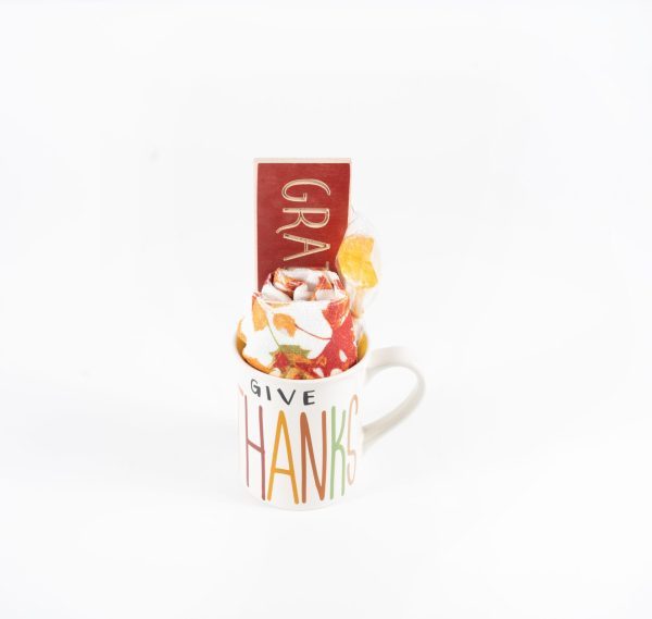 A white "Give Thanks" mug, decorated with colorful letters, holds a rolled-up autumn-themed cloth, a red packet labeled "GRATITUDE," and other small items partially visible. This charming piece could be part of an inviting gift basket or paired with a free food certificate for an extra touch of warmth (Product Name: Give Thanks Mug Gift Basket with Free Food Certificate).