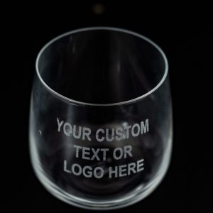 A clear, empty glass from the Pair of Rocks/Juice Glasses collection with the text "YOUR CUSTOM TEXT OR LOGO HERE" etched in white on its surface. The glass, ideal for juice, is set against a dark background.