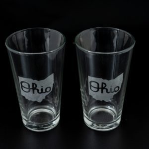 Two clear pint glasses from the "Pair of Pint Glasses - State of Ohio with Script Ohio" set are placed side by side against a black background. Each glass features an outline of the state of Ohio with the word "Ohio" written in a cursive script inside the outline.