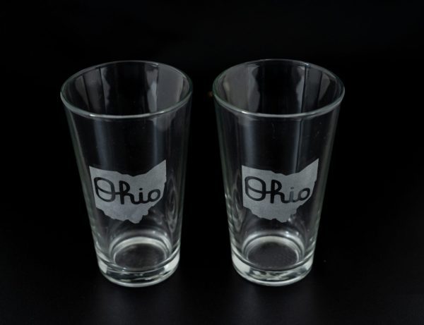 Two clear pint glasses from the "Pair of Pint Glasses - State of Ohio with Script Ohio" set are placed side by side against a black background. Each glass features an outline of the state of Ohio with the word "Ohio" written in a cursive script inside the outline.