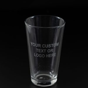 A clear glass from the "Pair of Pint Glasses - Your Custom Text/Logo Here" set is depicted against a black background, with the text "YOUR CUSTOM TEXT OR LOGO HERE" etched onto its surface. The empty glass stands upright, perfectly centered in the image. This elegant piece is ideal for displaying your custom text or logo and can be paired with another pint glass for added impact.