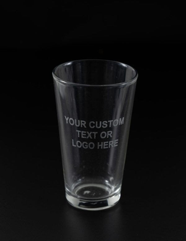 A clear glass from the "Pair of Pint Glasses - Your Custom Text/Logo Here" set is depicted against a black background, with the text "YOUR CUSTOM TEXT OR LOGO HERE" etched onto its surface. The empty glass stands upright, perfectly centered in the image. This elegant piece is ideal for displaying your custom text or logo and can be paired with another pint glass for added impact.