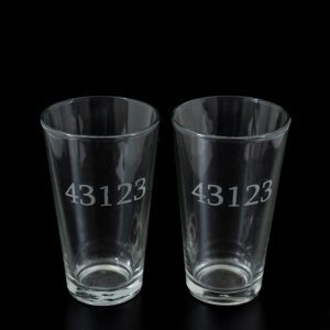 Two clear pint glasses, engraved with the number "43123" resembling unique zip codes, are showcased against a black background. Identical in shape and size, this pair of pint glasses is placed side by side.