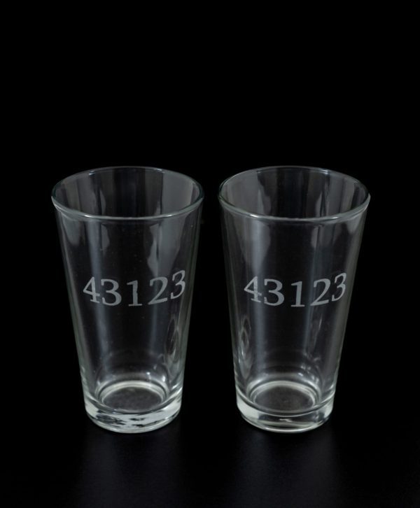 Two clear pint glasses, engraved with the number "43123" resembling unique zip codes, are showcased against a black background. Identical in shape and size, this pair of pint glasses is placed side by side.