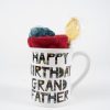 A white Happy Birthday Grandfather Mug with Free Food Certificate displays "Happy Birthday Grandfather" in colorful, patterned letters. The gift pack inside the mug includes a red and green knit item and a yellow, round, wrapped candy, all set against a plain white background.