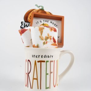 A white ceramic mug with the word "Grateful" in colorful letters and "Incredibly" above in black letters. This Grateful Mug Gift Basket with Free Food Certificate includes a rolled-up festive towel adorned with autumn leaf patterns, a small decorative frame, a mini pumpkin ornament, and a Free Food Certificate.